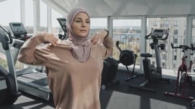 Güzel Müslüman kadın tesettüre ve spor kıyafetlerine bürünmüş, spor salonuna gitmeden önce ısınırken omuz hareketi yapıyor ve egzersiz yapıyor.