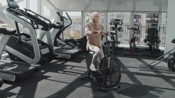 ヒジャーブのムスリム女性とジムでのトレーニング中に静止運動自転車に乗るスポーツウェア — ストック動画