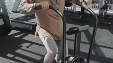 Spor kıyafetleri ve tesettürlü genç Müslüman bir kadının spor salonunda antrenman yaparken kırtasiye bisikleti sürüşünü çek.