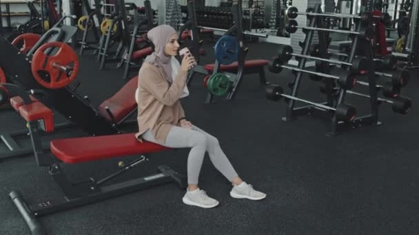 穆斯林妇女穿着头巾和运动服坐在体育馆的长椅上 锻炼后喝水 — 图库视频影像