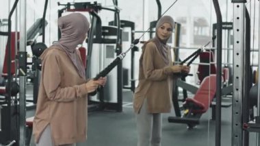 Başörtüsü ve spor kıyafetleri içinde Müslüman bir kadın kablo makinesiyle dikilip aynaya bakıyor.