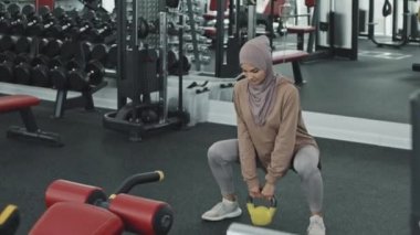 Spor giysileri ve tesettürlü Müslüman bir kadın elinde kettlebell, spor salonunda antrenman yaparken çömeliyor.