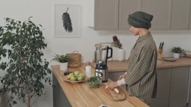 Başörtülü Müslüman kadın evde mutfakta smoothie pişirirken muz kesip miksere koyuyor.