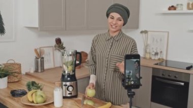 Kafiyeli güzel Müslüman kadın evde mutfakta yemek videosu çekerken meyveyi gösteriyor ve akıllı telefon kamerasıyla konuşuyor.