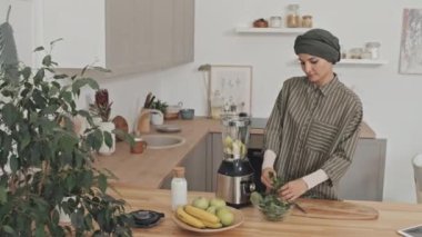 Başörtülü güzel Müslüman kadın evde meyve püresi yaparken ıspanak yapraklarını karıştırıcıya koyup süt ekliyor.