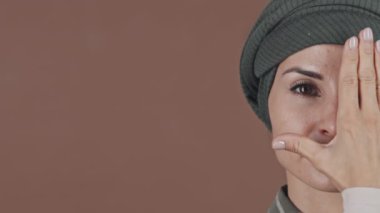 Başörtülü genç Müslüman kadının stüdyo portresi. Yüzünün yarısını eliyle kapatıyor ve kameraya bakıyor.