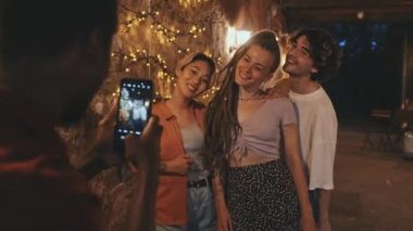 Orta halli, çoklu etnik kökenli arkadaş grubu yazın günlük kıyafetlerini giyip geceleri dışarıda fotoğraf çekiyorlar. Sarı ışıklarla süslenmiş duvara karşı poz veriyorlar.