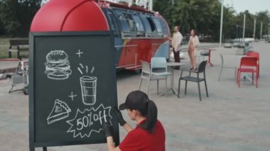Üniformalı genç kadın yemek kamyonu işçisi güneşli bir günde dışarıdaki tahtaya özel bir teklifte bulunurken meslektaşı arka plandaki insanlara sokak yemeği satıyor.