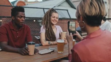 Genç bir kadının, kız arkadaşının işaret levhasını gösterirken, bira içerken ve yaz akşamları açık çatı katındaki kafede eğlenirken orta boy fotoğrafı.