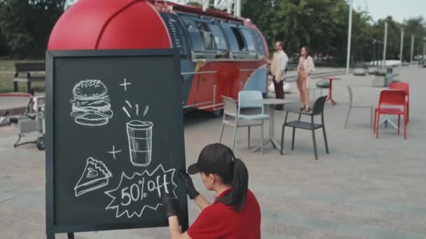 晴れた日に外の黒板に特別なオファーを描く制服を着た若い女性食品トラック労働者彼女の同僚が背景に人々にストリートフードを販売しながら — ストック動画
