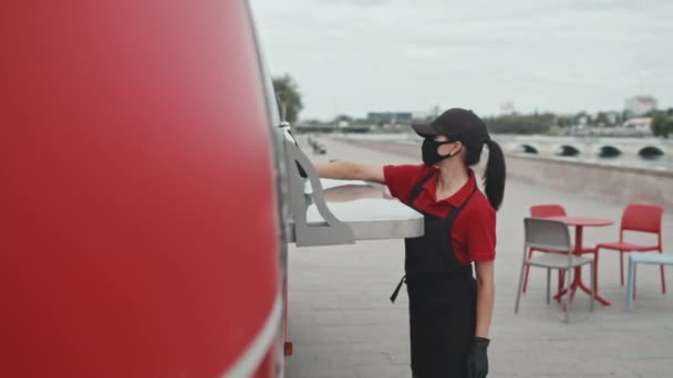顔のマスクでストリートフードの若い女性の売り手の中ショットと均一なクリーニング赤いトラックと開く前に布でカウンター外 — ストック動画