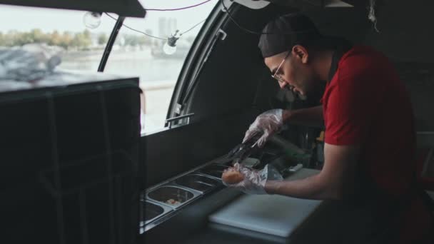 Střední záběr mladého muže ze Středního východu v uniformě, jak dělá hot dog v jídelním voze, bere ingredience z kovových nádob