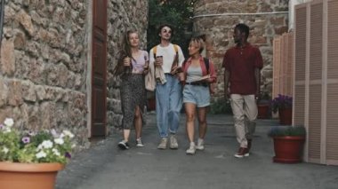Sıradan giyimli dört genç arkadaşın dar tuğla yolda yürürken ve yaz gününü birlikte sohbet ederken tam boy yavaş çekimleri.