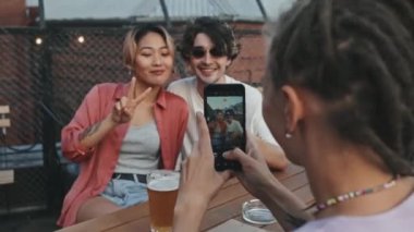 Rastalı genç bir kadının akıllı telefonuyla arkadaşlarının fotoğrafını çekerken çatıdaki barda oturup bira içerken ve yaz akşamının tadını çıkarırken orta boy bir fotoğrafı.
