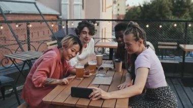 Dört farklı genç arkadaşın akıllı telefondan video görüşmesi yaparken, konuşurken ve kameraya el sallarken, yaz akşamları çatıdaki barda, ahşap masada otururken.