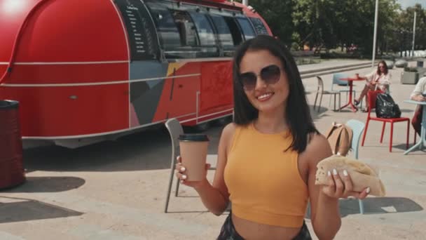 背景に赤いフードトラックと夏の屋外でタコと紙コーヒーカップと立ってカメラで笑顔サングラスの若い美しい女性の中肖像画 — ストック動画
