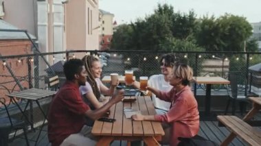 Orta halli, çoklu etnik çeşitlilikte, bira bardaklarıyla kadeh kaldıran erkek ve kadın arkadaşlarının yaz akşamları sarı ışıklarla süslenmiş çatıdaki kafede yan yana oturduğu bir fotoğraf.