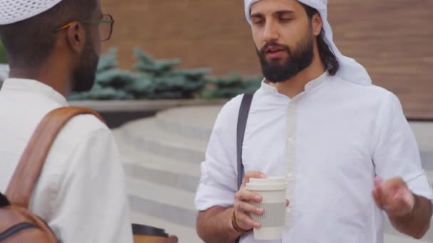 髭を生やした男が白いシャツとスカーフを着てコーヒーカップを持って街の外で黒人男性の友人と話している — ストック動画