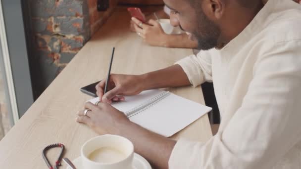 Mediano Hombre Negro Sonriendo Pensando Bebiendo Capuchino Escribiendo Cuaderno Sentado — Vídeo de stock