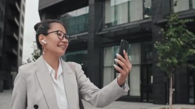 Resmi giyinmiş, gözlüklü, neşeli Asyalı iş kadınının şehirde yürürken ve cep telefonundan biriyle sohbet ederken çekilmiş bir fotoğrafı.
