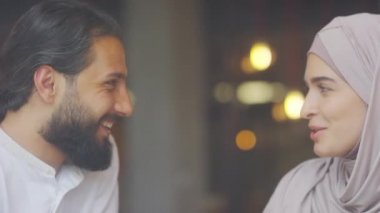 Kafkasyalı, kestane rengi gözlü, tesettürlü Müslüman bir kadının omuzları kalkık, kapalı arka planda iki yüzlü sakallı bir adamla konuşuyor.