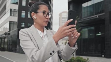 Asyalı iş kadınının resmi kıyafetleri ve gözlükleriyle dışarıda dikilip cep telefonuyla konuşurken çekilen görüntüsü.