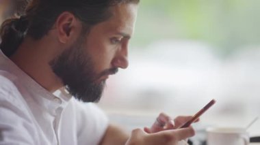 Beyaz gömlek giyen, akıllı telefon kullanan, gündüz vakti pencerenin yanında oturan sakallı, çift cinsiyetli adamın yan görüntüsü.