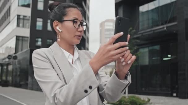 外に立って 携帯電話でビデオ通話で話している正式な摩耗や眼鏡でアジアのビジネス女性の追跡ショット — ストック動画