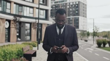 Takım elbiseli, kravatlı ve gözlüklü Afro-Amerikan işadamının caddede yürürken cep telefonuyla yazarken çekilmiş bir fotoğrafı.