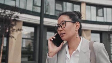 Resmi giyinmiş, gözlüklü, neşeli Asyalı iş kadınının sokakta yürürken cep telefonuyla konuştuğu yakın plan çekimleri.