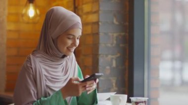 Kafkasyalı, tesettürlü, modern kafede akıllı telefon kullanan, pencerenin kenarında oturan genç Müslüman kadının orta boy yakın çekimi.