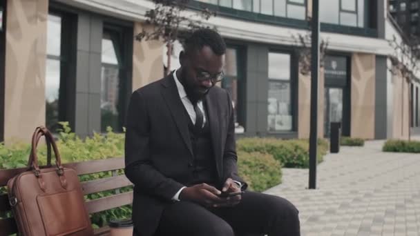 정장을 안경을 아프리카 미국인 사업가가 벤치에 전화로 타이핑하는 모습을 포착하는 — 비디오