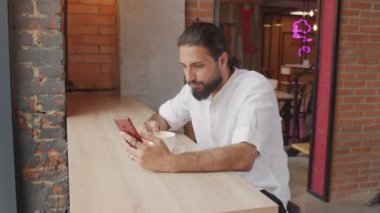 Orta boylu, sakallı, beyaz gömlek giyen, latte içen, modern kahve dükkanında pencere kenarında oturan, gündüz vakti akıllı telefon kullanan iki yüzlü genç adam.