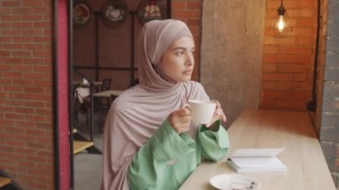 Kafasında tesettür olan, kahve fincanı tutan, gündüzleri modern kafede oturan, kameraya bakıp gülümseyen fındık gözlü beyaz kadının yavaş çekimde portresi.