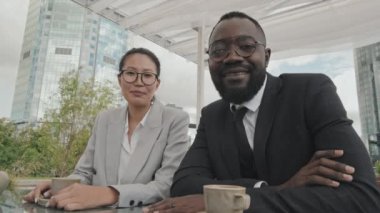 Asyalı iş kadını ve resmi giyinmiş Afro-Amerikalı iş adamı POV açık kafedeki masada oturmuş kahve içerken kamerayla konuşuyor.