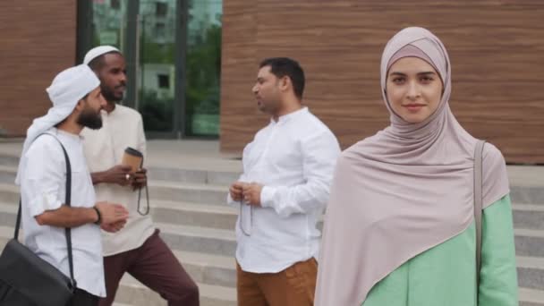 美しい白人ムスリム女性のウエストアップヒジャーブを身に着けています 笑顔とカメラを見て オフィスビルで話している3人の男性の前で立って — ストック動画
