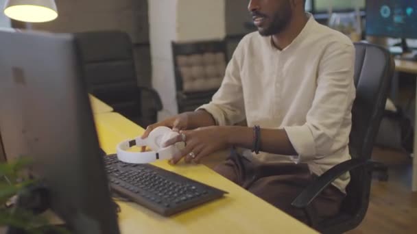 クフィと白いシャツを着た黒人イスラム教徒の男が机の上に座って 昼間に耳の上のヘッドフォンをして コンピュータを使って — ストック動画