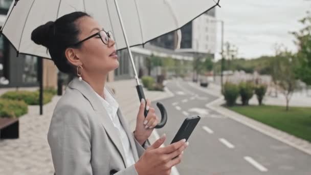 市内の歩道に立ちながら 傘を持って携帯電話に入力し 正式な摩耗でエレガントなアジアの実業家のショットを追跡 — ストック動画