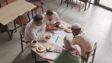 Modern kafede masada oturan, evrak işleri yapan, iş görüşmeleri yapan, konuşan dört Müslüman erkek ve kadının üst düzey görüşü.