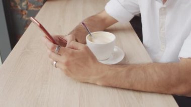 Beyaz gömlek giyen, latte içen, kahve dükkanında pencere kenarına oturmuş, akıllı telefon kullanan kırpık adamların yakın çekimleri.