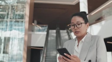 Asyalı iş kadınının resmi kıyafetleri ve gözlükleri ile ofis lobisinde yürürken ve cep telefonuyla yazarken görüntüsü.