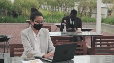 Asya 'lı iş kadını ve Afro-Amerikan iş adamının açık kafede maske takıp salgın sırasında uzaktan çalışmalarını gösteren görüntüler.