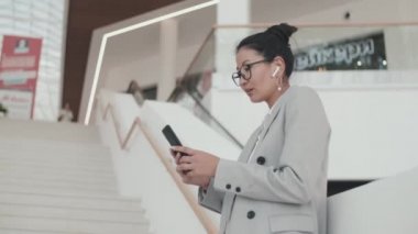 Asyalı iş kadınının resmi giysileri ve gözlükleriyle alışveriş merkezindeki merdivenlerde durup cep telefonuyla yazarken orta boy görüntüsü.