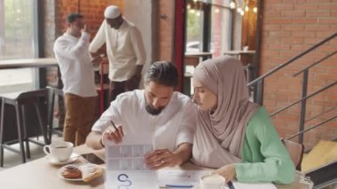 Kafkasyalı, tesettürlü, sakallı, melez bir adamın modern kafede oturmuş evrak işleriyle uğraşan, konuşan, arka planda dikilen insanların orta boy yakın çekimi.