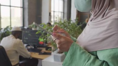 Yan görünüş ela gözlü beyaz kadın tesettür ve yüz maskesi takıyor, tablet bilgisayar kullanıyor, gündüzleri ofiste duruyor.