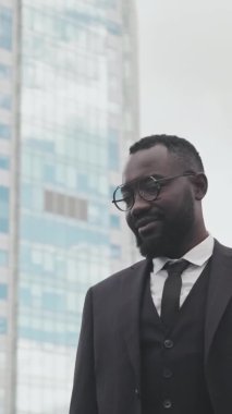 Üç parçalı takım elbiseli, kameraya gülümseyen ve şehirde poz veren mutlu Afro-Amerikan işadamının el yapımı dikey portresi. Arkaplanda bina