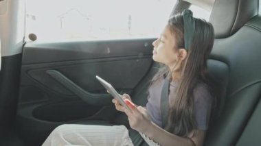 Arabanın arka koltuğunda otururken gülümseyen ve tablet kullanan 9 yaşındaki bir kızın el kamerası görüntüsü.
