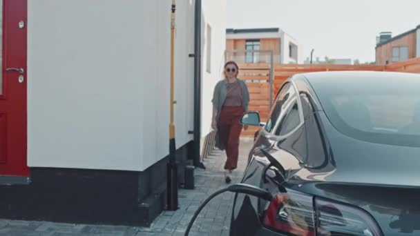 Tracking shot of stylish young woman in sunglasses unplugging electric car from charger outside her suburban house
