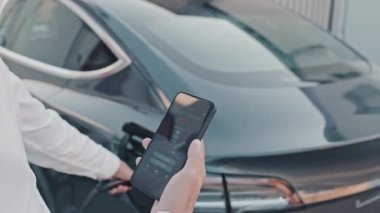 Cep telefonu tutan tanınmayan erkek sürücünün ellerini kapatın ve elektrikli arabasını şarj ederken ekrandaki uygulamaya bakın.