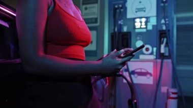 Geceleri EV istasyonunda elektrikli arabasına yaslanırken tanınmayan Afro-Amerikan bir kadının cep telefonu kullanırken orta bölüm görüntüsü.